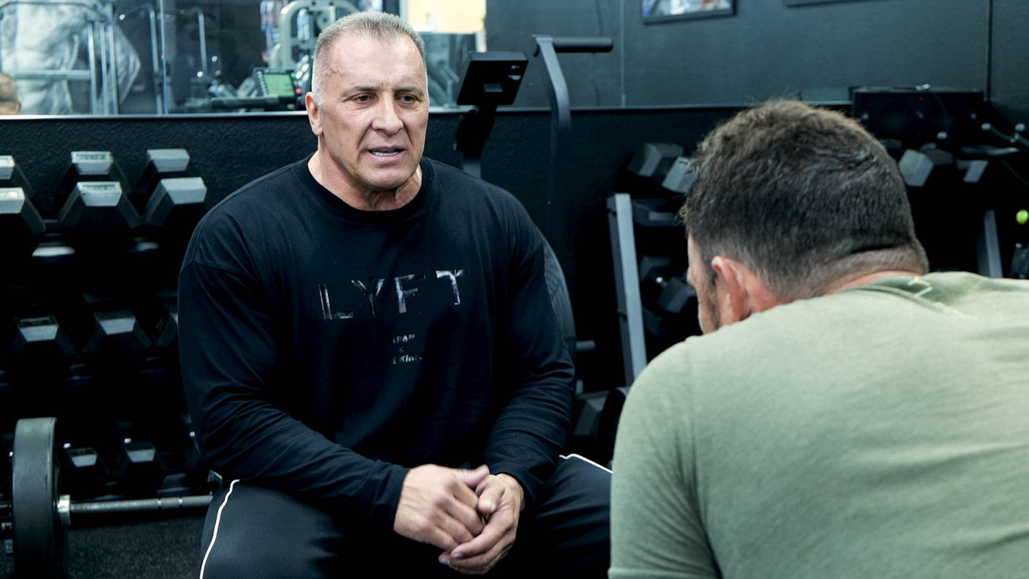 Milos Sarcev in black LYFT shirt talks with JYM Supps president beside dumbbell rack in gym during filmed discussion.