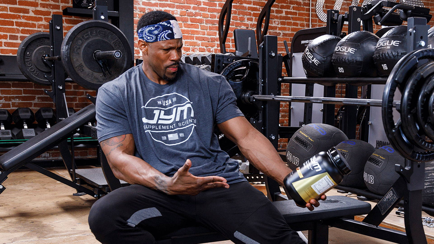 IFBB Pro Michael Norwood in JYM tee and bandana pouring a Pro JYM protein tub while seated on a gym bench by squat racks.