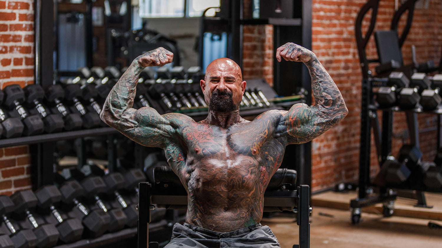Jim Stoppani hitting a double biceps pose on a bench in front of dumbbell racks, illustrating "Six Tips to Sick Arms".