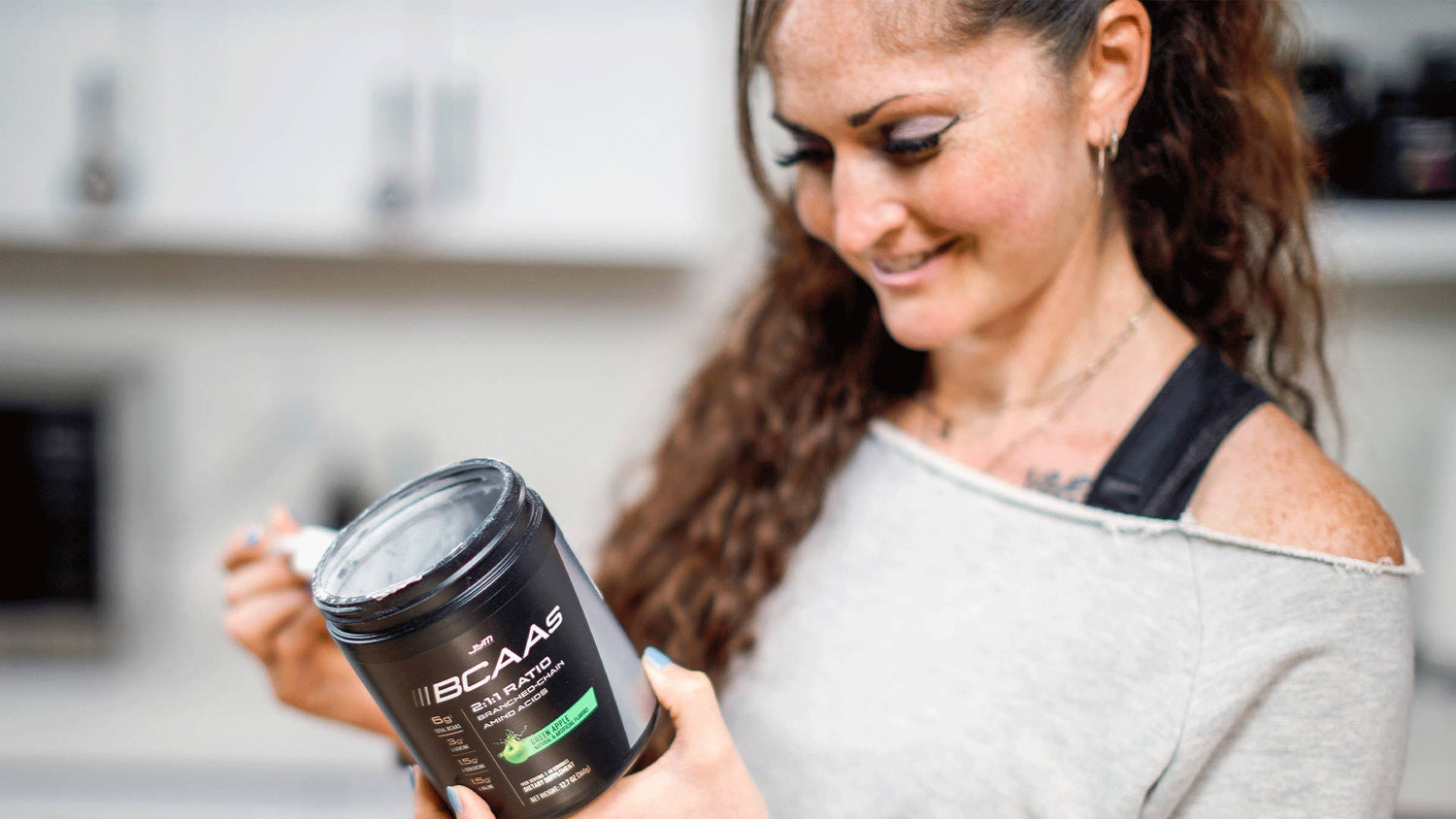 Smiling woman in gray top holding opened black tub of JYM BCAAs Green Apple in a kitchen, article BCAAs Pre Workout Tips.