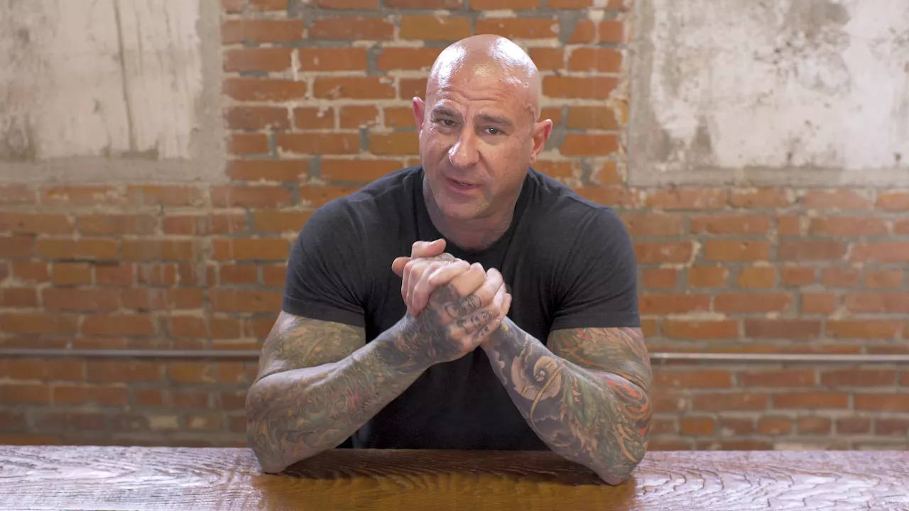 Jim Stoppani with tattooed arms clasped at a wooden table against a brick wall, discussing "Is Your Whey Protein Spiked?"