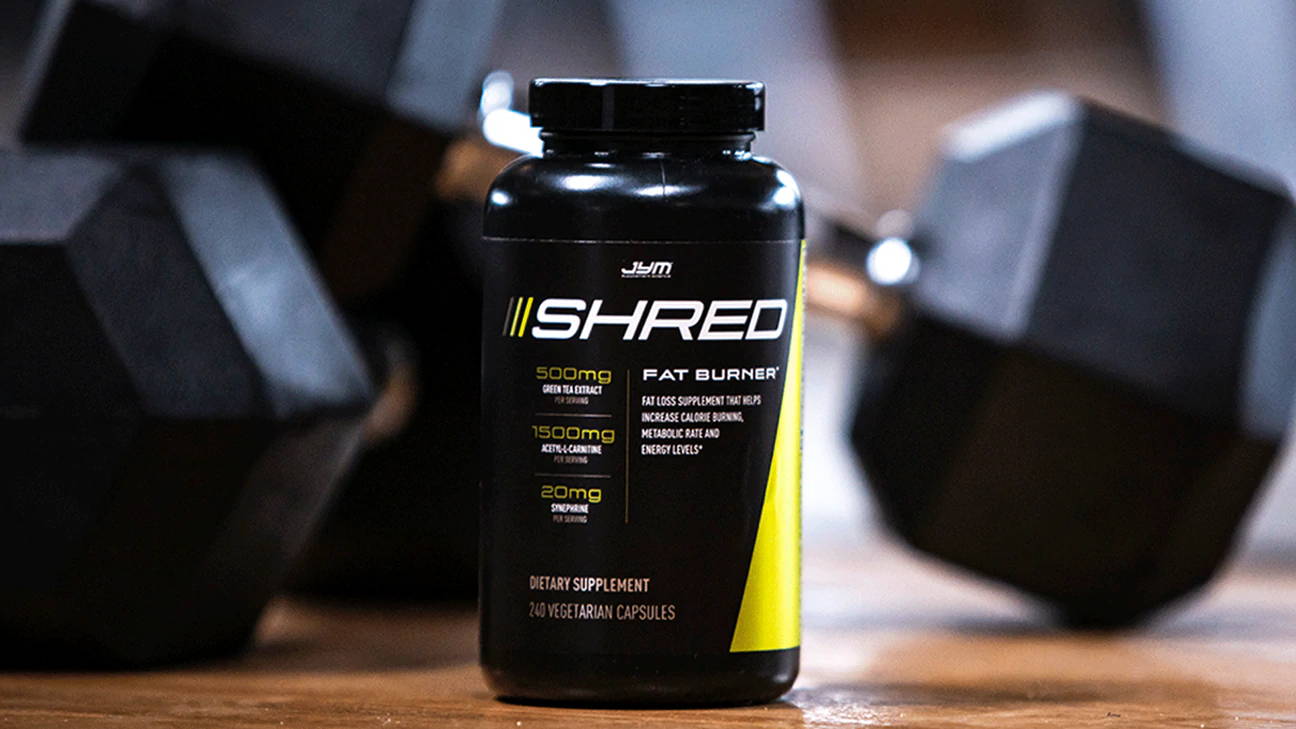 Black Shred JYM Fat Burner bottle with yellow accents and 240 capsules, sitting on wood floor beside black hex dumbbells.