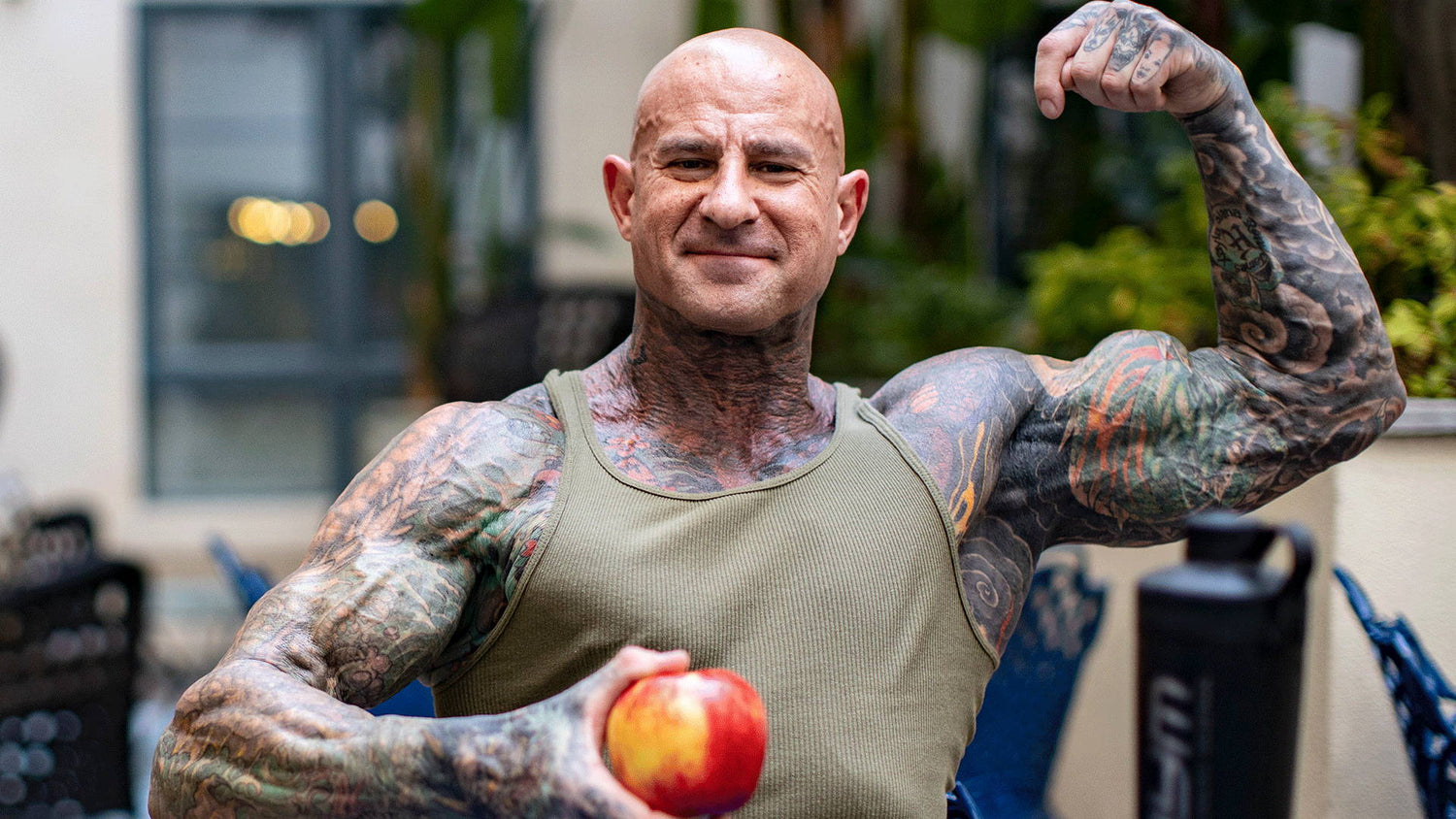 Jim Stoppani in olive tank top flexing bicep and holding a red apple outdoors, illustrating The 5 Best Pre Workout Foods.