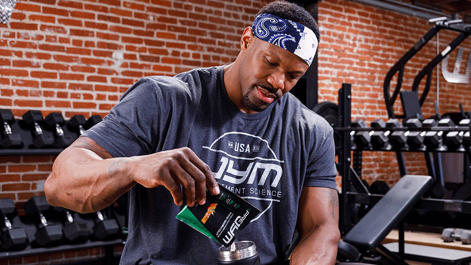 Athlete in gray JYM tee tearing open Pre JYM single-serve packet over shaker cup, dumbbell rack behind — 5 Supplements article.
