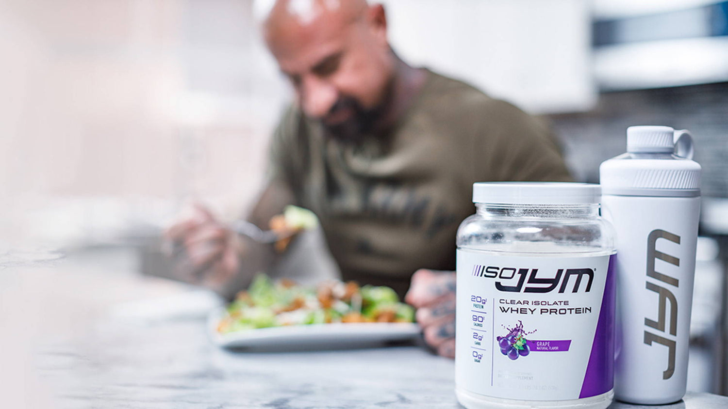 Iso JYM grape clear whey isolate container and JYM shaker on kitchen counter, tattooed man blurred behind eating salad.