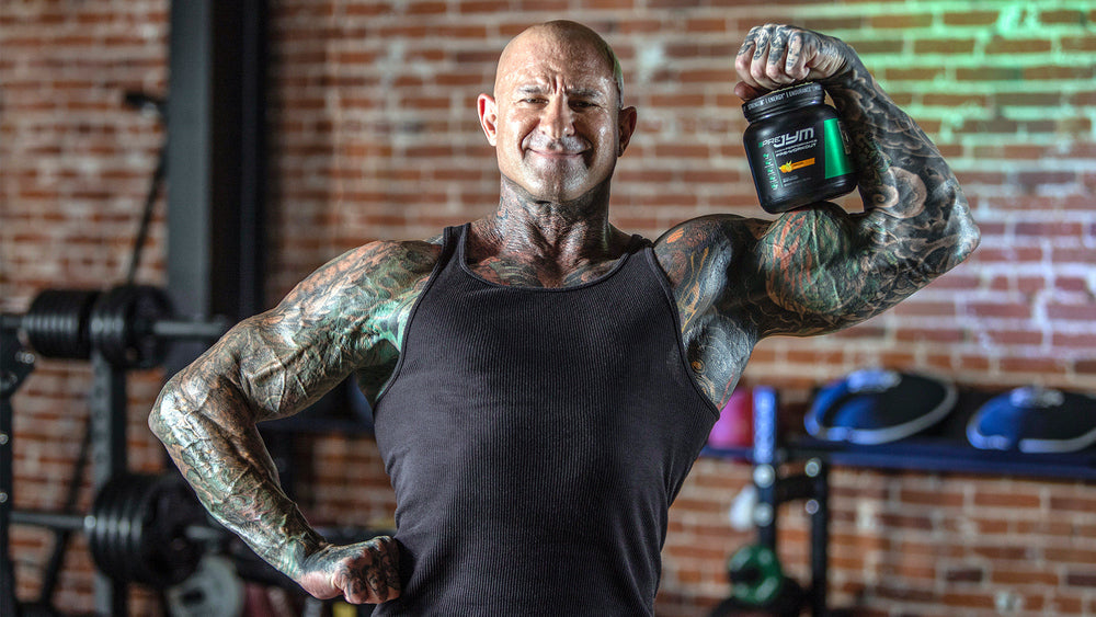 Jim Stoppani smiling and flexing bicep beside Pre JYM tub, brick gym wall behind, for Why Pre JYM is NOT Just a Pre-Workout.