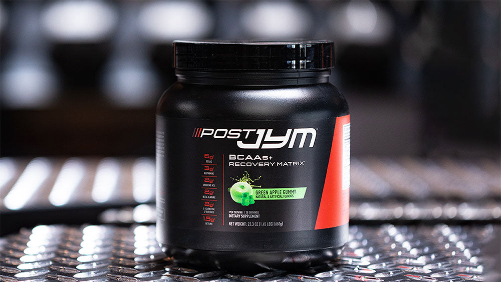 Post JYM BCAAs+ Recovery Matrix tub, Green Apple Gummy flavor, displayed on metal gym flooring with dumbbell rack behind.