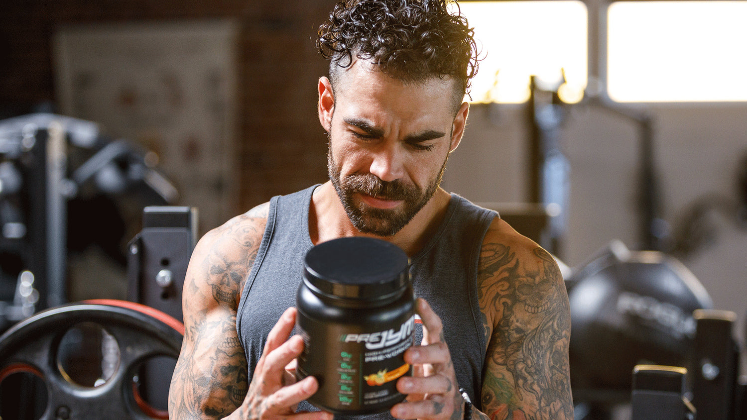 Tattooed man in gray tank top holding and reading a black Pre JYM pre-workout tub in a gym, for "Why You NEED Pre JYM".