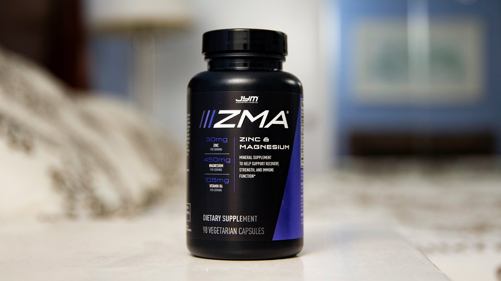 Black ZMA JYM bottle with purple label showing zinc and magnesium dosages, sitting on a nightstand in a bedroom setting.